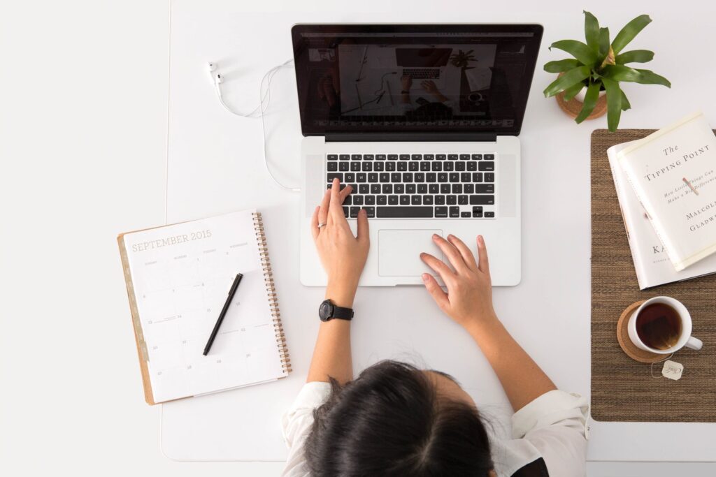 A picture taken from the top looking down of a woman typing on a laptop, a calendar and coffee cup sitting beside her. 