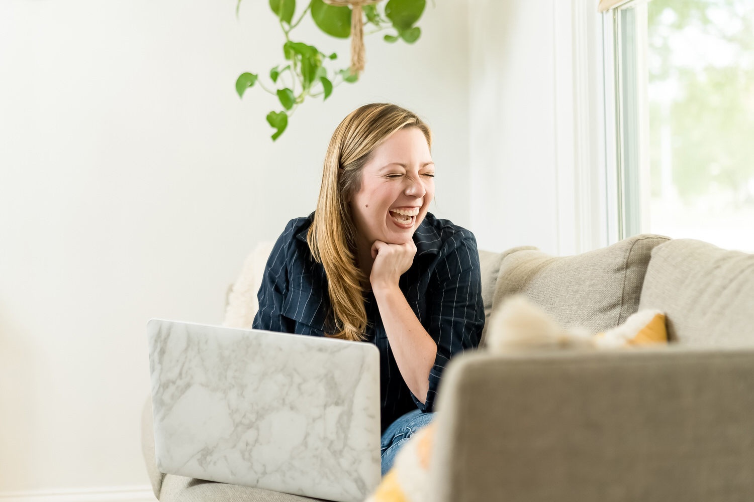 A candid picture of a woman sitting on a couch working on a laptop and laughing. 