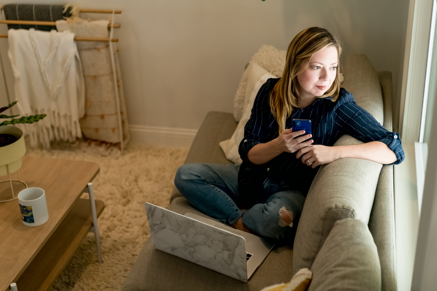 A picture of a woman in a blue shirt sitting on a couch, staring out a window, er phone in her hand. 