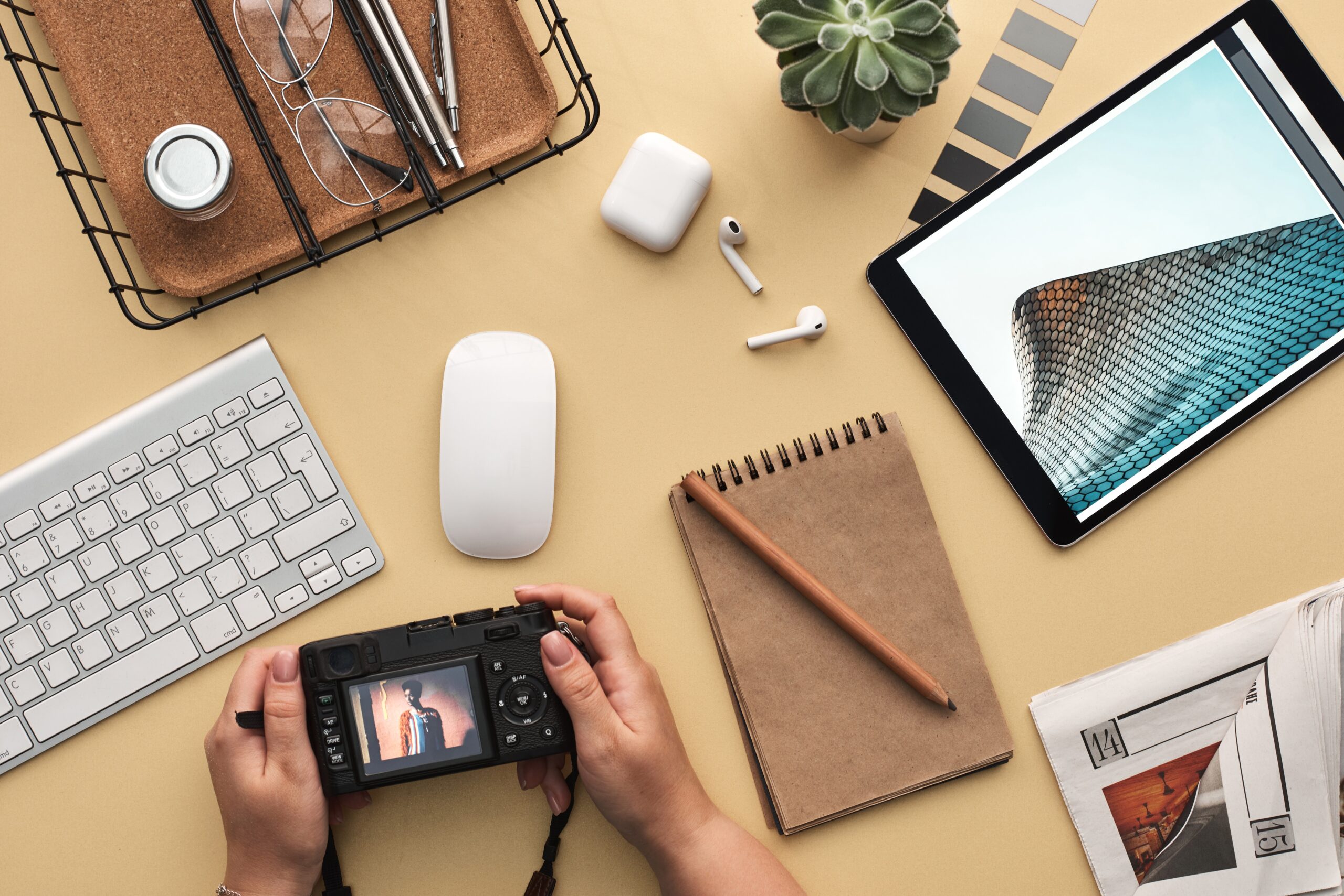 A picture of a pair of hands holding a camera, and other desktop accessories sitting on a neutral table top. 